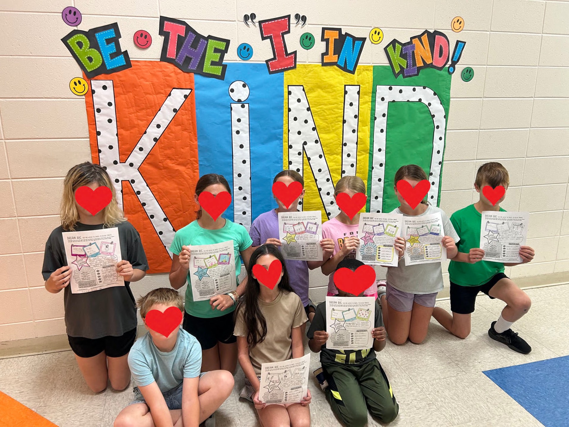 Students at Opp Elementary in Alabama holding their completed Glitter for a Cause kindness plans in front of a 'Be the I in Kind' classroom display