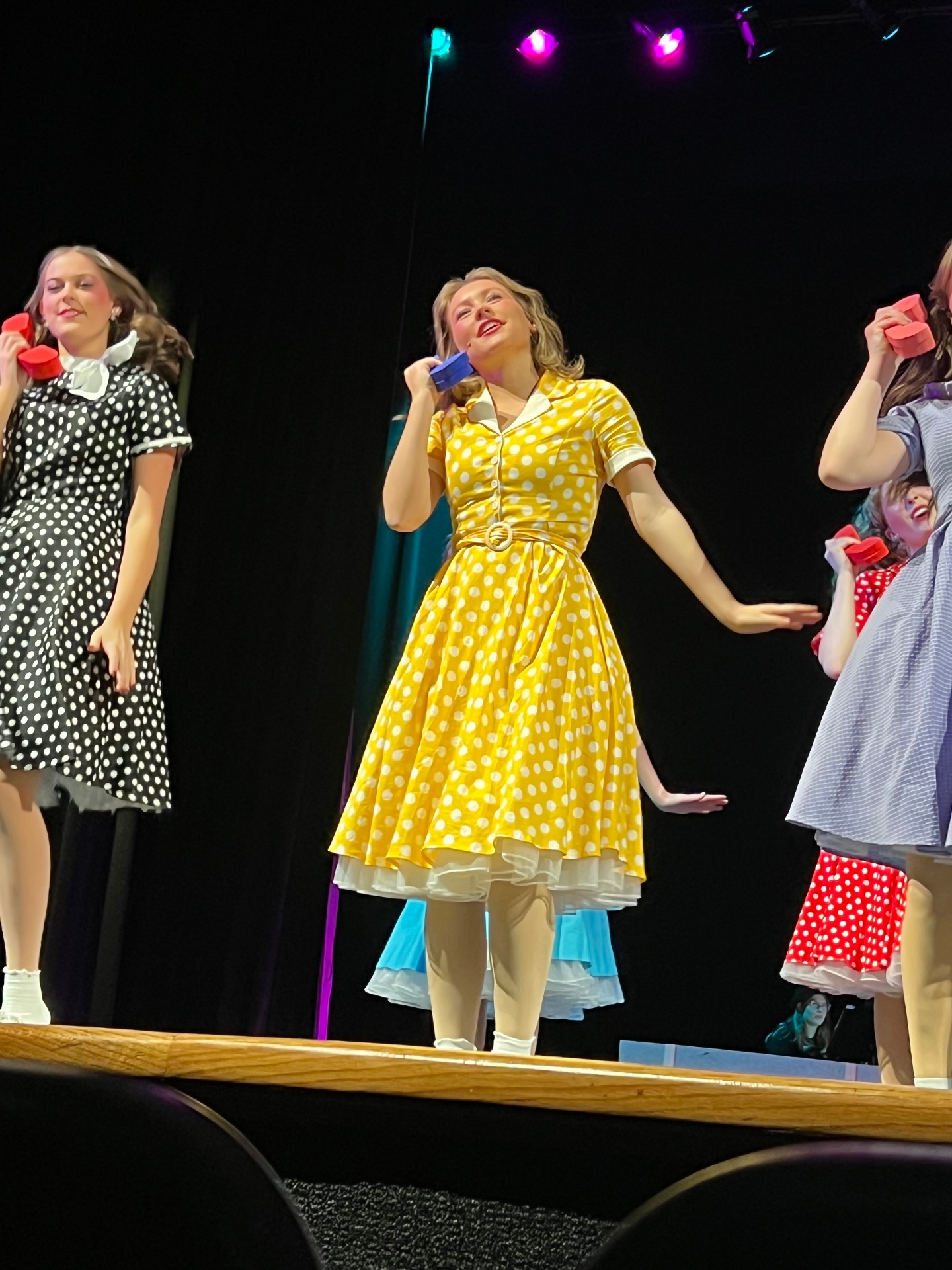 Callie Chapman center stage in a yellow polka-dot dress holding a purple prop phone during Mountain Brook High School's production of Bye Bye Birdie