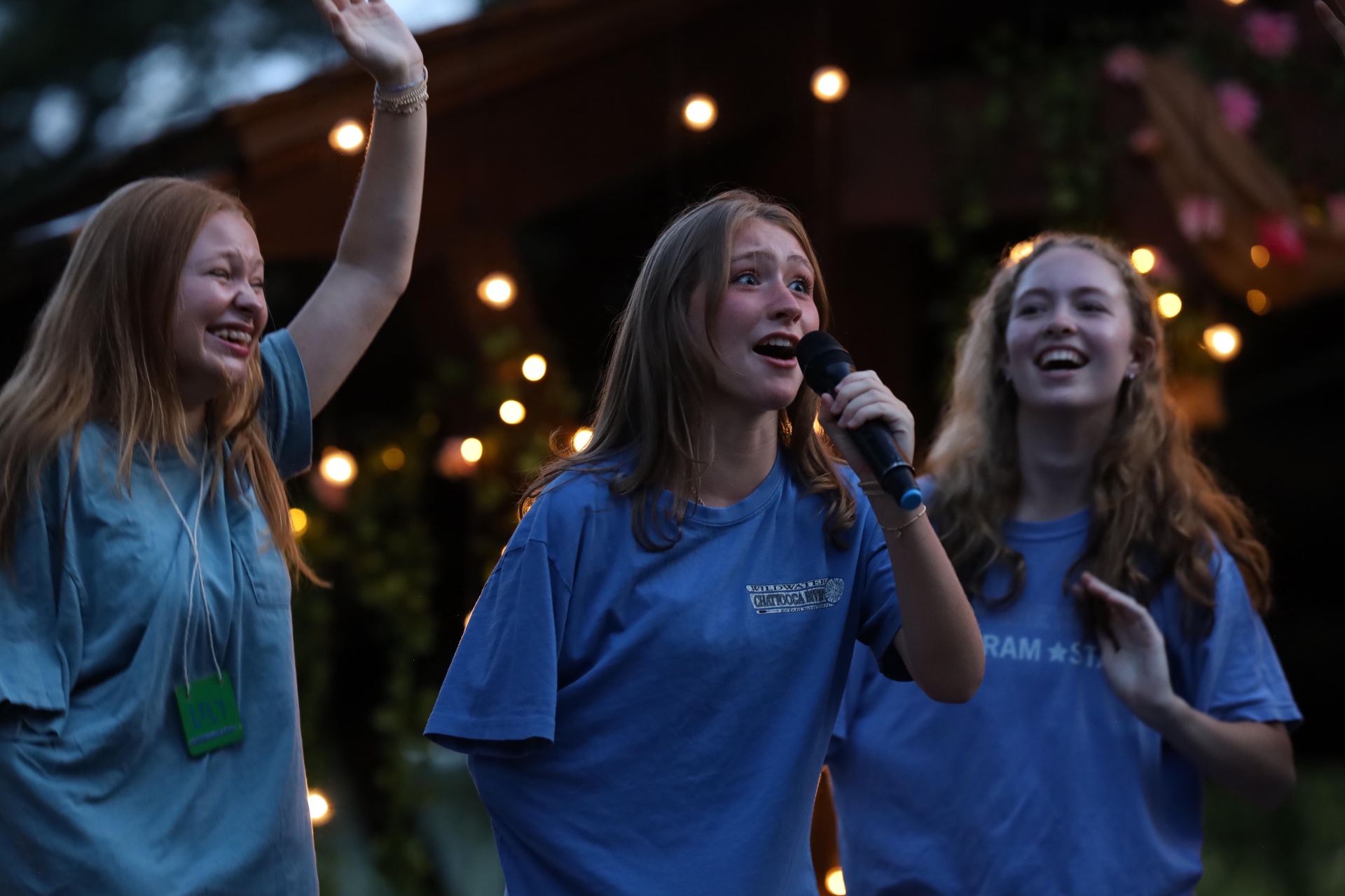 Callie Chapman singing into a microphone at Greystone Camp for Girls with two friends cheering beside her under string lights
